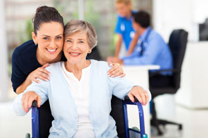 A female Nurse is standing behind an elderly female patient who is sitting in a wheelchair. They are in a hospital hallway smiling.